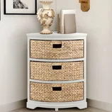 A corner chest with three woven drawers and a white top, decorated with a vase and books. It is placed beside framed art and a wall-mounted sconce.