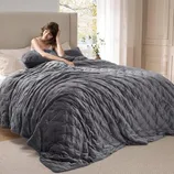 A gray velvet weighted blanket is draped over a bed with a person sitting up, holding a matching pillow.