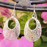 Silver earrings with a floral cut-out design, featuring an oval shape and hook attachments, set against a backdrop of pink flowers.
