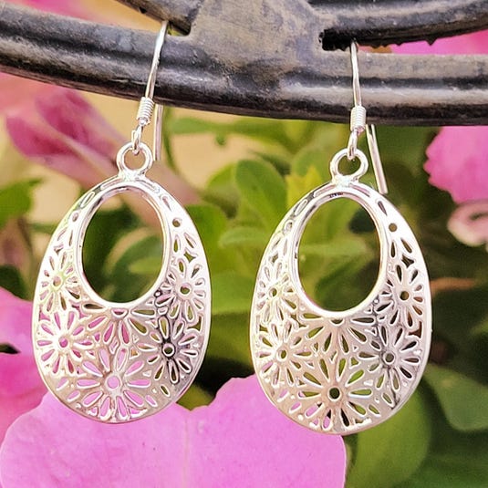 Silver earrings with a floral cut-out design, featuring an oval shape and hook attachments, set against a backdrop of pink flowers.
