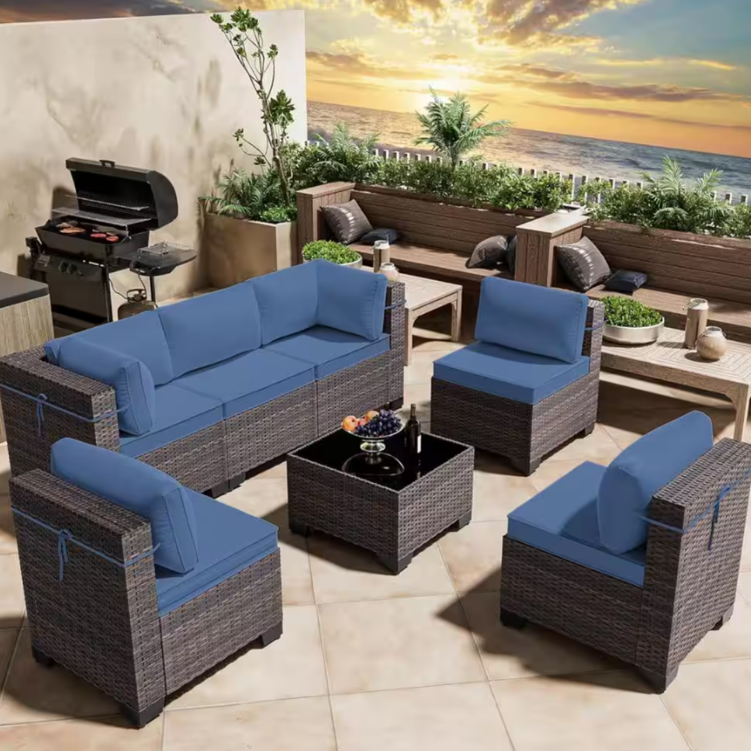 7-piece outdoor sectional set with blue cushions includes a three-seater sofa, two armchairs, two corner chairs, a central coffee table, and an ottoman. The furniture features a wicker design and is arranged on a patio with a view.