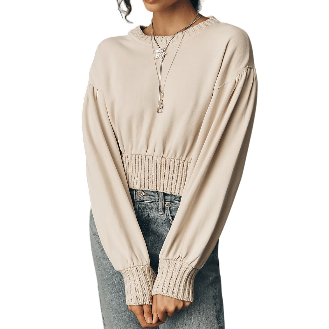 A woman is wearing a light beige sweater with ribbed cuffs and waistband, paired with high-waisted blue jeans. She is also wearing a long pendant necklace.