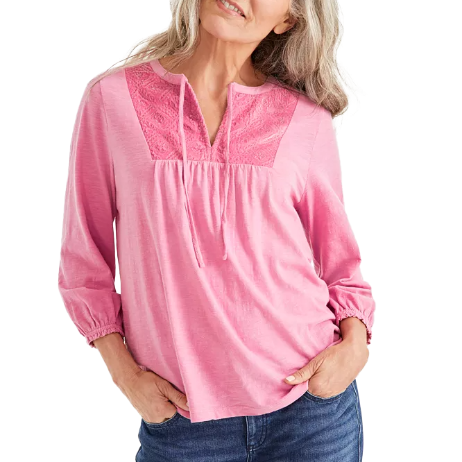 A pink Velvet-Bib Peasant Top features three-quarter sleeves with elastic cuffs, a delicate velvet bib with embroidery, and a split neckline with tassel ties. It’s paired with jeans, offering a casual yet stylish look.