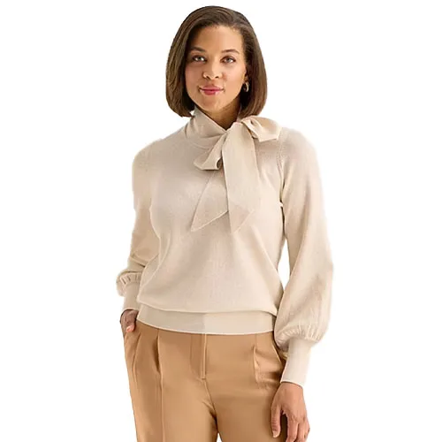 A woman is wearing a light-colored bow-tie sweater paired with beige trousers, standing against a white background.