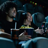 Two people are enjoying popcorn and drinks in a movie theater. The popcorn bucket has Regal branding, and the drink cup features a Pepsi logo.