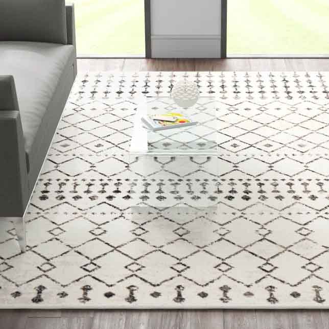 5' x 7' Southwestern area rug with a cream base featuring a geometric diamond pattern in dark tones, placed near a gray sofa and a clear glass table on wooden flooring.