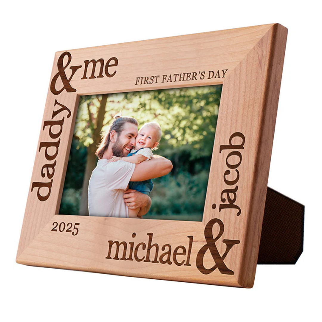 A wooden photo frame engraved with \“daddy & me,\“ \“First Father's Day,\“ \“michael & jacob,\“ and the year \“2025,\“ featuring a photo of a man holding a baby outdoors.