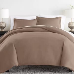 A brown bedding set featuring a duvet cover and matching pillow shams, complemented by white bed sheets. Each side of the bed has a lamp with a round, textured base.