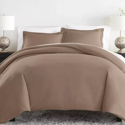 A brown bedding set featuring a duvet cover and matching pillow shams, complemented by white bed sheets. Each side of the bed has a lamp with a round, textured base.