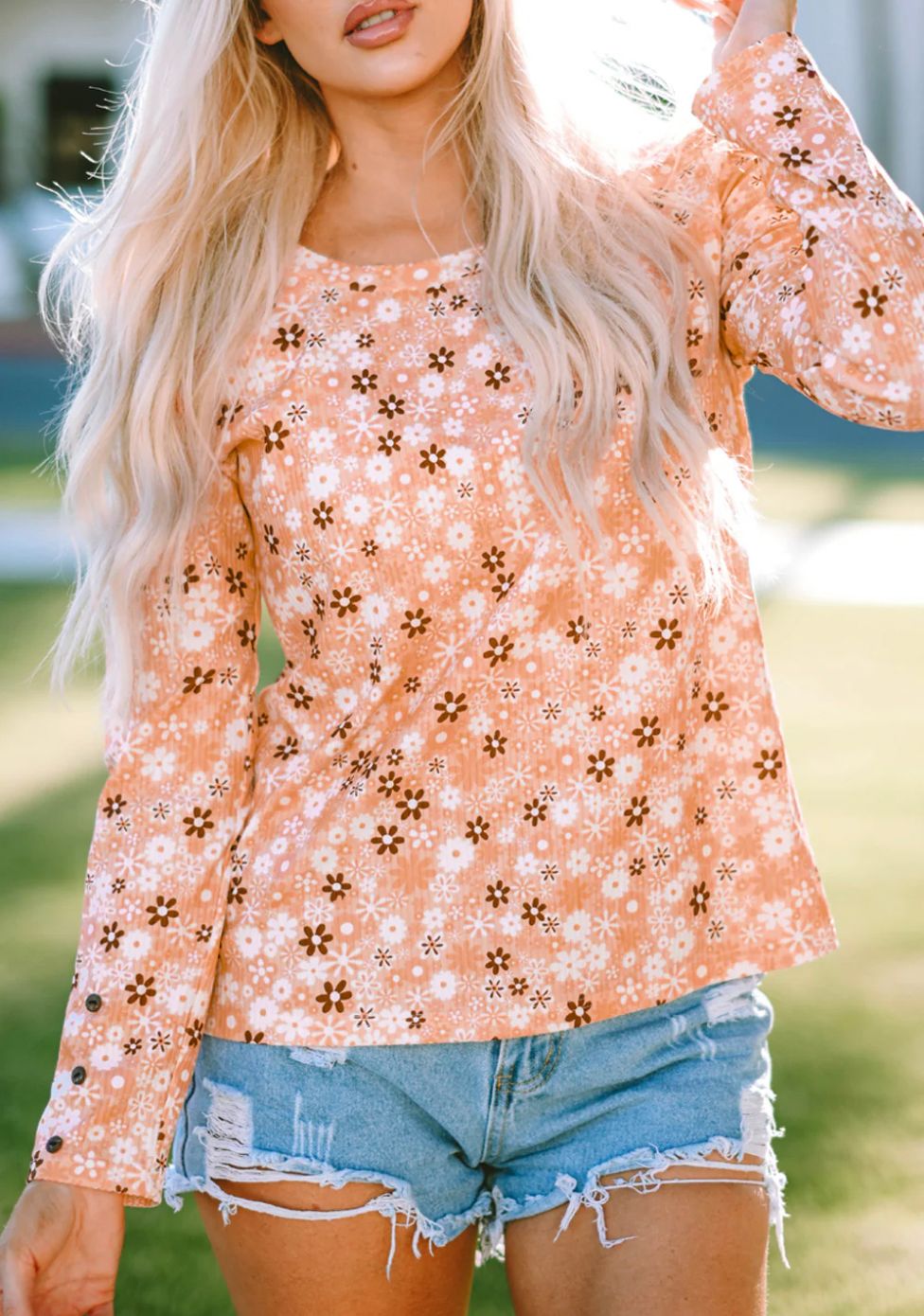 A woman is wearing a long-sleeved floral knit top in pastel peach tones with denim shorts.