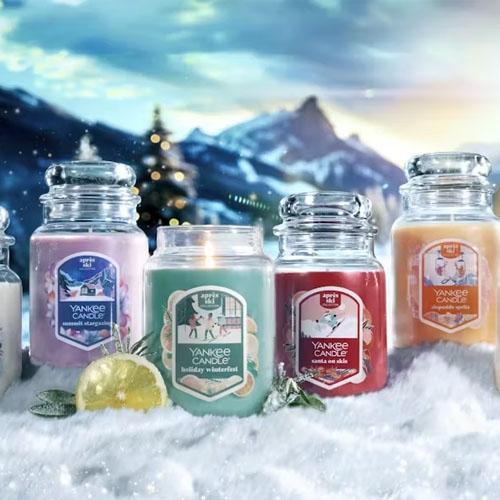 A collection of Yankee Candle jars on a snowy surface, featuring scents like \“Evergreen Mist,\“ \“Alpine Morning,\“ \“Holiday Hearth,\“ \“Candlelit Cabin,\“ and \“Pomegranate Gin Fizz,\“ set against a winter mountain backdrop.