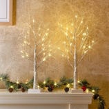 Lighted birch tree decorations with warm white lights are placed on a mantel alongside green pine garland and pine cones.