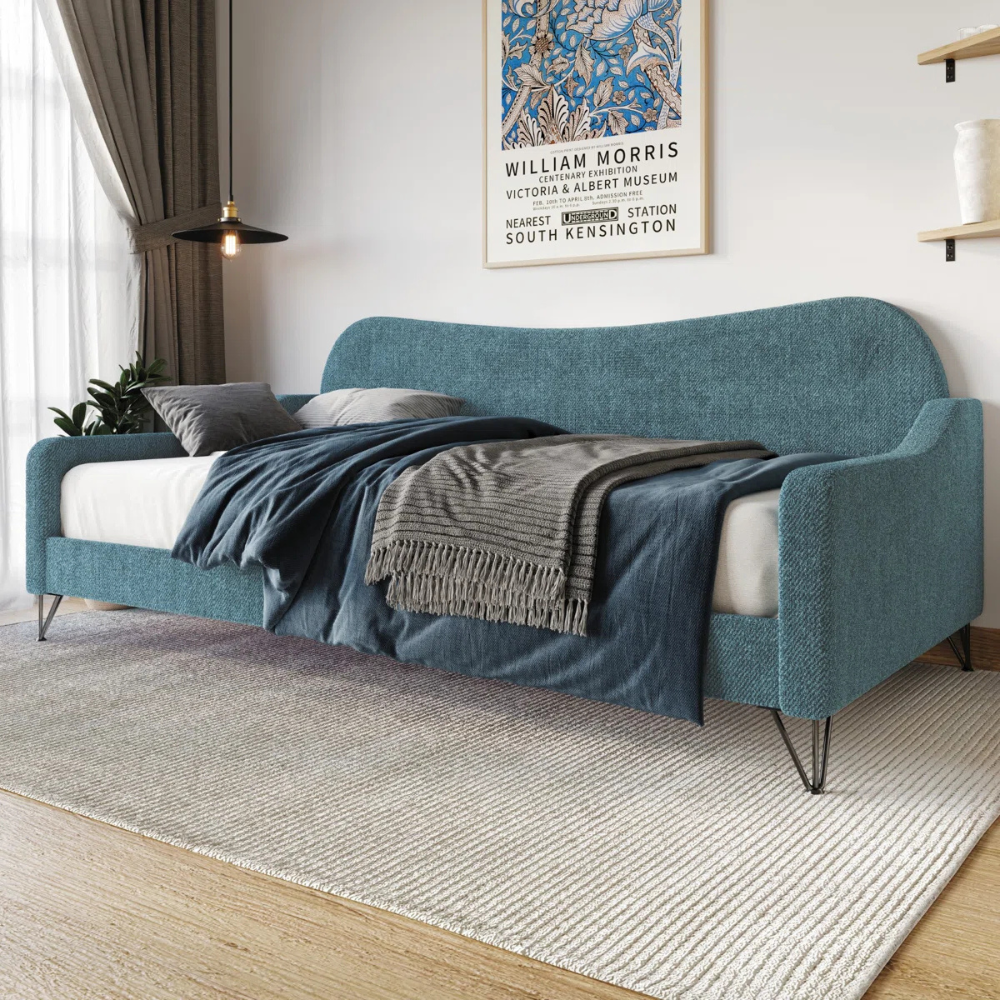 A blue upholstered daybed with metal legs is adorned with gray and navy bedding. Nearby are a curtain, a plant, and a framed William Morris poster on the wall.