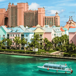 A turquoise boat moves through clear water, with colorful waterfront buildings and a large hotel complex with distinct architecture in the background, surrounded by palm trees.