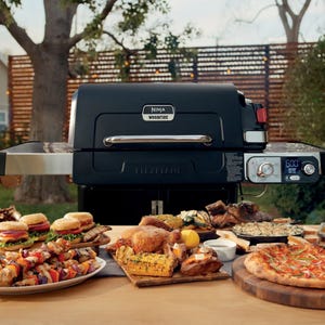 A Ninja Woodfire Flexflame grill is surrounded by a variety of grilled foods, including burgers, kabobs, roast chicken, corn on the cob, and pizza, set on an outdoor table.