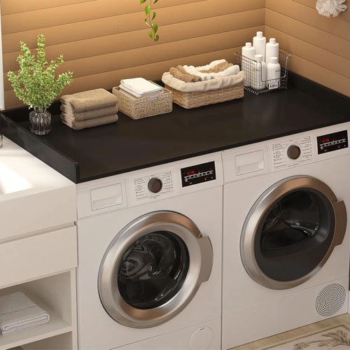 A laundry setup featuring a front-loading washing machine and dryer beneath a black countertop, with towels, a plant, and toiletries neatly arranged on top.