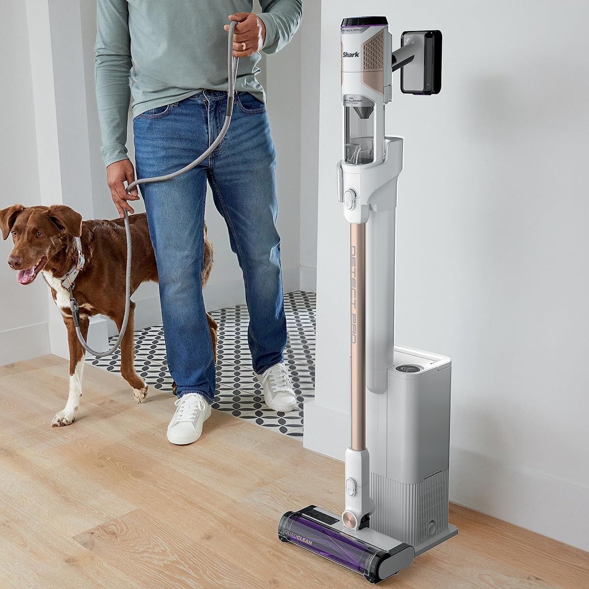 A cordless stick vacuum cleaner docked in its charging station, with a person holding a dog on a leash nearby.