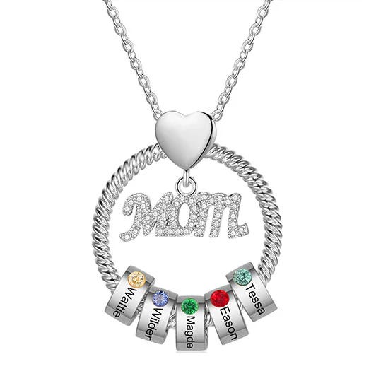 A necklace with a heart-shaped pendant holding a circular ring. Inside the ring is the word \“MOM\“ in ornate letters, accompanied by rings with colored gemstones and engraved names: Mattie, Wilder, Maggie, Eason, Tessa.