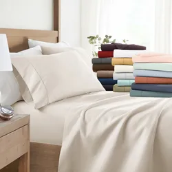 A bed is outfitted with a neutral-colored deep-pocket fitted sheet and matching pillowcases; additional folded sheet sets in various colors are displayed at the foot of the bed.