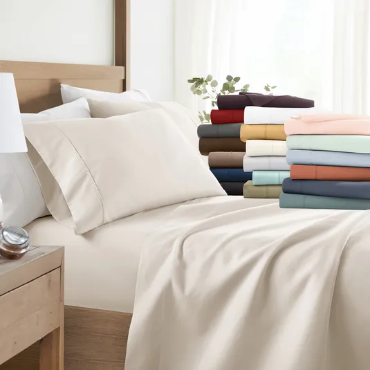 A bed is outfitted with a neutral-colored deep-pocket fitted sheet and matching pillowcases; additional folded sheet sets in various colors are displayed at the foot of the bed.