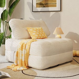 A cozy beige armchair with ribbed upholstery is styled with a yellow floral pillow and matching throw. There's a small lamp, a potted plant, and an open book nearby, set against a neutral rug and wall art.