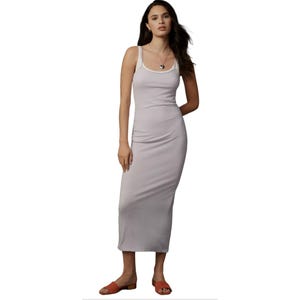 A woman is wearing a sleeveless, form-fitting, ankle-length gray dress paired with orange sandals. She has a necklace and earrings as accessories.