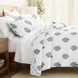A neatly made bed features white bedding with blue floral patterns, accompanied by matching pillows, set in a bright room with wooden furniture and a leafy plant.