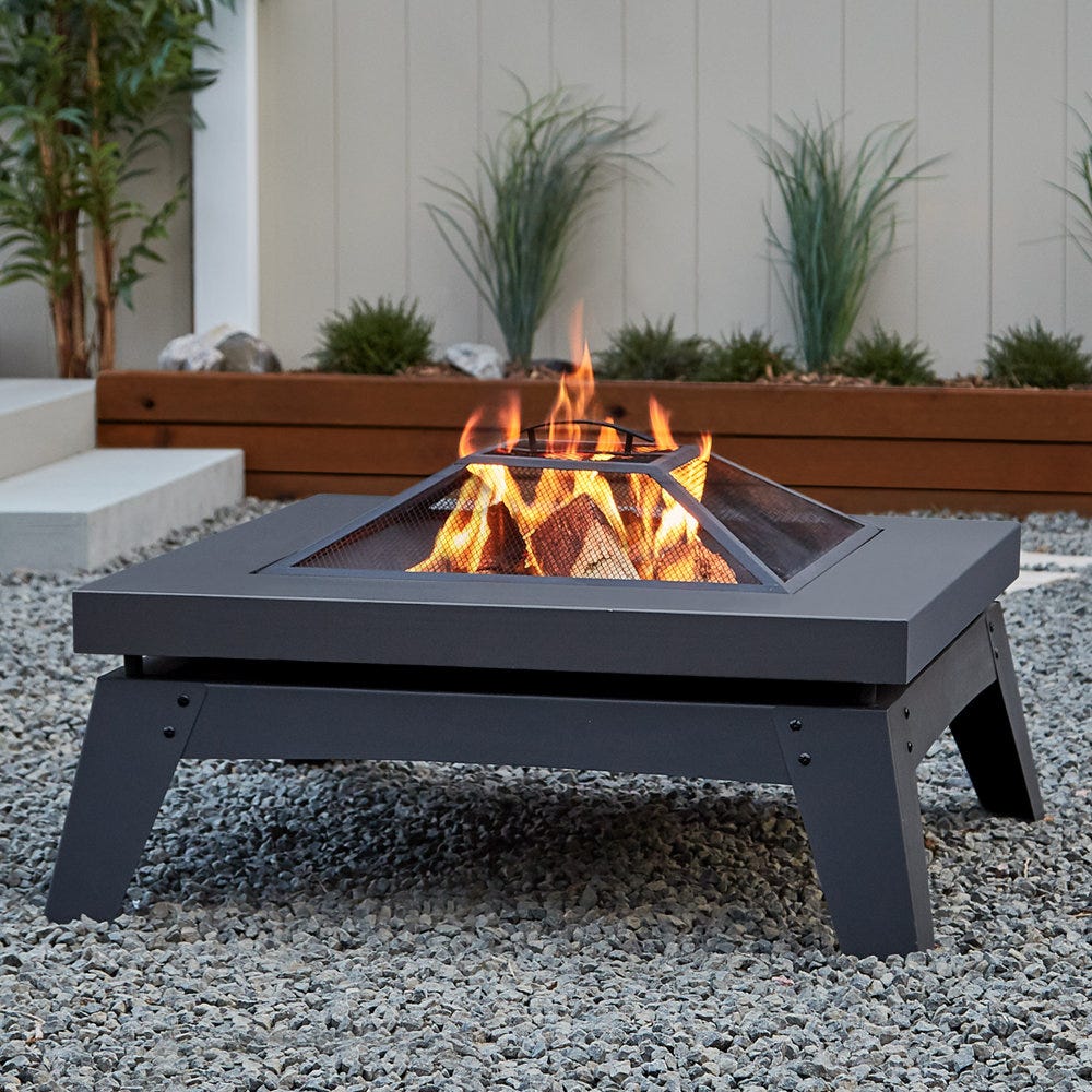 A square wood-burning fire pit with a mesh lid is centered on a bed of gravel, emitting a warm flame. The pit has a low-profile, modern design with black metal construction.