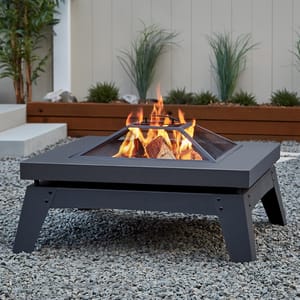 A square wood-burning fire pit with a mesh lid is centered on a bed of gravel, emitting a warm flame. The pit has a low-profile, modern design with black metal construction.