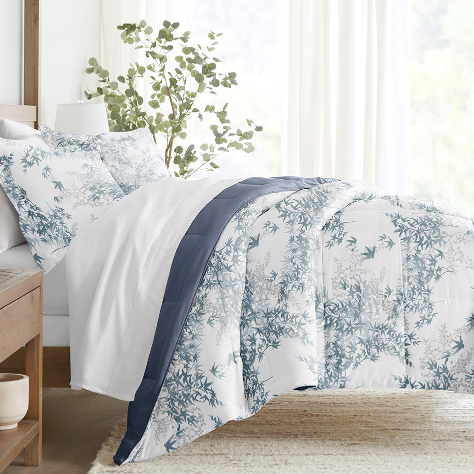 A bed is adorned with a white comforter featuring a blue floral pattern, a dark blue stripe, and matching pillowcases. A wooden side table and a plant are visible nearby, creating a tranquil bedroom setting.