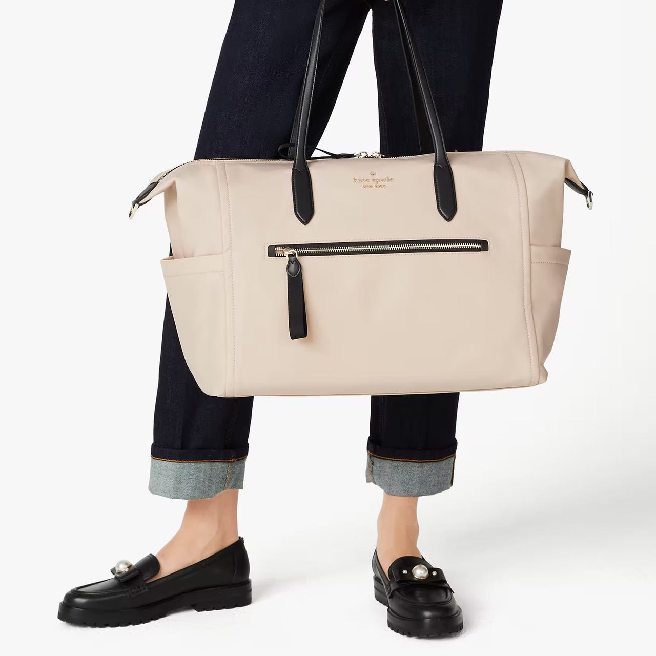 A person holding a large beige tote bag with a front zipper pocket, paired with dark loafers and cuffed jeans.