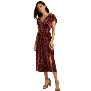 A woman is wearing a burgundy velvet midi dress with short sleeves, floral pattern, pleated detailing, and a side slit. She pairs it with open-toe heeled sandals.