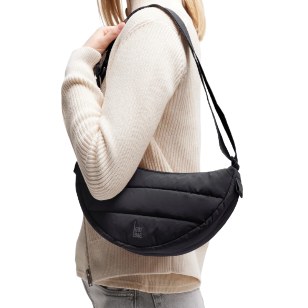 A person is wearing a black, crescent-shaped shoulder bag from Got Bag made of quilted material, paired with a light, ribbed sweater and light-colored pants.