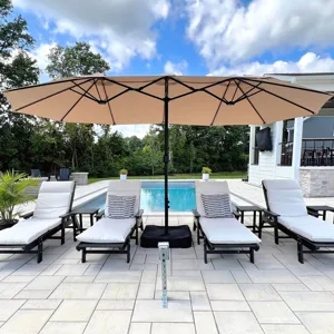 A large patio umbrella covers cream-colored lounge chairs with striped cushions by a pool, surrounded by a paved patio and greenery.