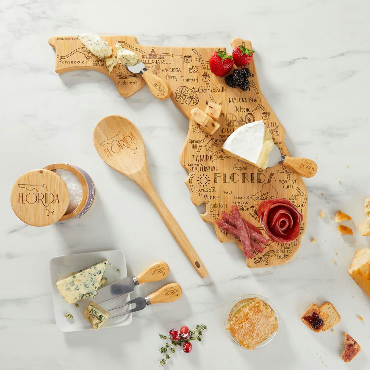 A Florida-shaped wooden cheese board displays various cheeses, meats, and berries. Accompanied by a wooden spoon, salt cellar, cheese spreaders, and a small dish of honeycomb, all branded with \“Florida.\“