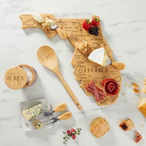 A Florida-shaped wooden cheese board displays various cheeses, meats, and berries. Accompanied by a wooden spoon, salt cellar, cheese spreaders, and a small dish of honeycomb, all branded with \“Florida.\“