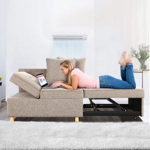A woman reclines on a multifunctional beige couch with an adjustable headrest and built-in black storage unit, using a laptop.