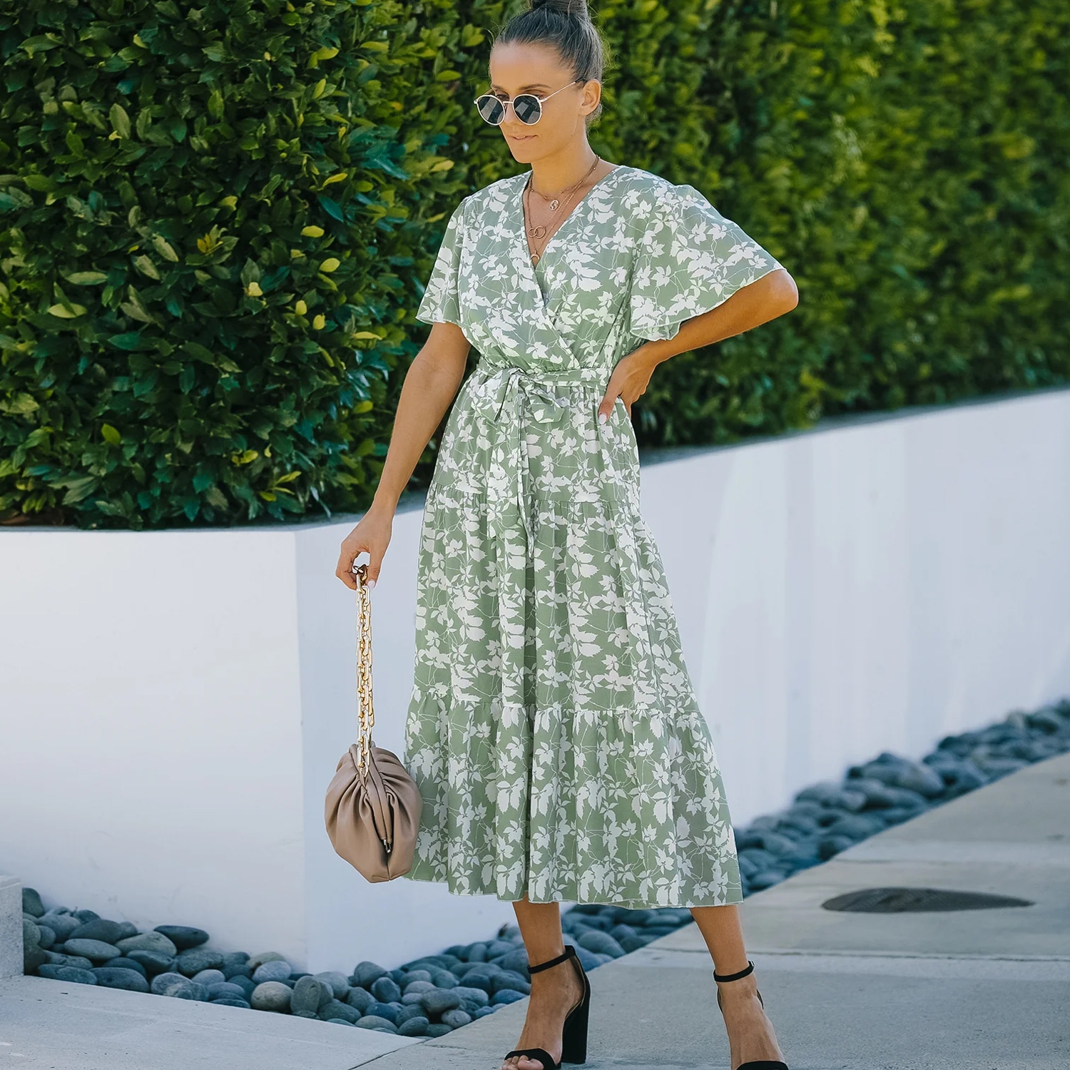 A woman in sunglasses wears a green floral wrap dress and black heeled sandals, holding a beige handbag with a chain strap, standing by a modern outdoor setting with lush greenery.