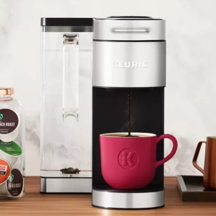 A Keurig coffee machine brewing into a pink mug is displayed on a counter next to a jar of assorted coffee pods, a small copper pitcher, and a white candle.