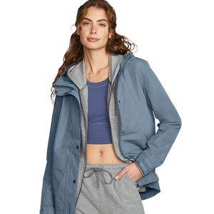 The Eddie Bauer Rainfoil Storm Jacket is a waterproof, breathable jacket with a blue outer shell. It features a hood and snap closures, worn here over a gray hoodie and paired with gray sweatpants and a blue crop top.
