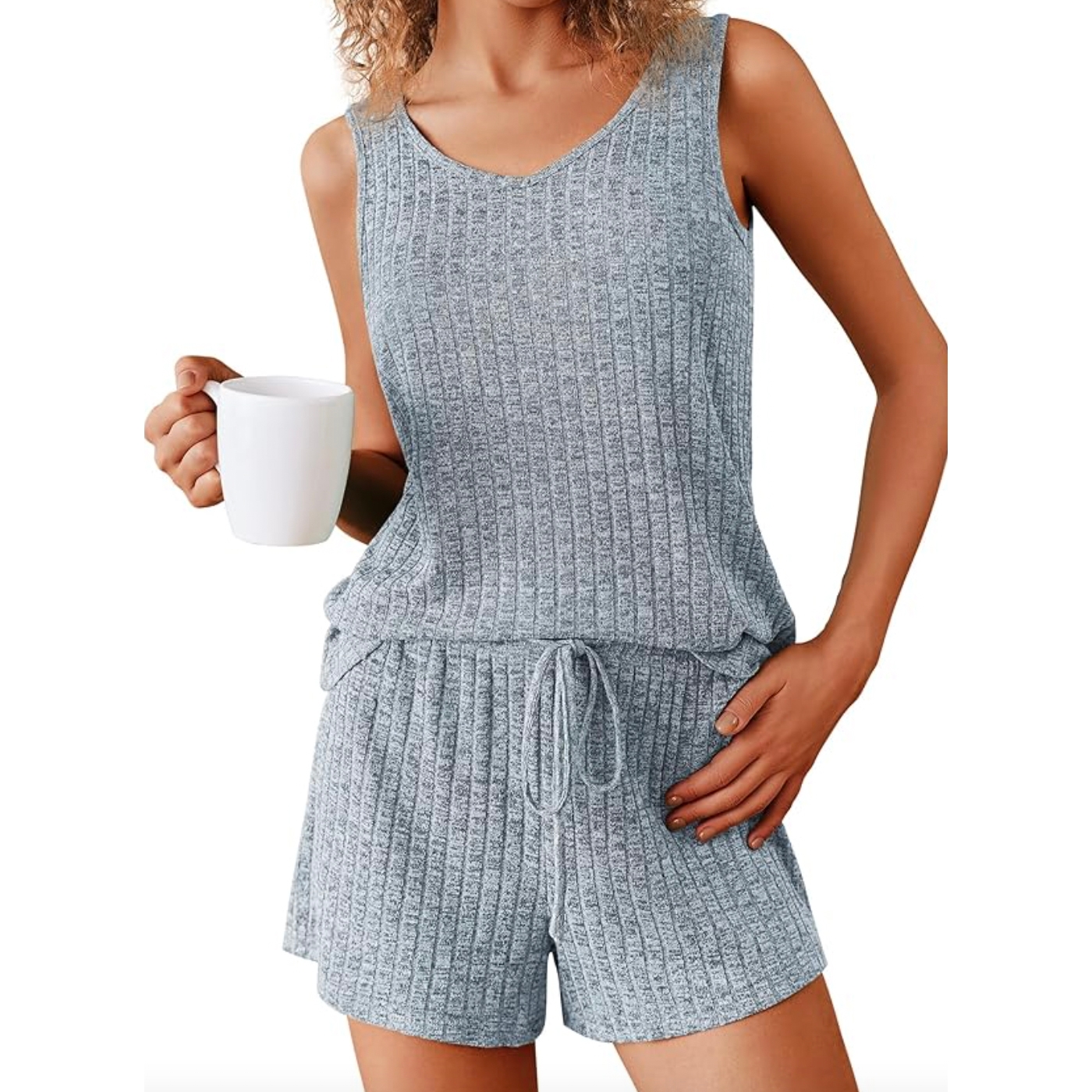 A woman is wearing a sleeveless, light gray, ribbed-knit tank top and matching drawstring shorts, holding a white mug in hand.