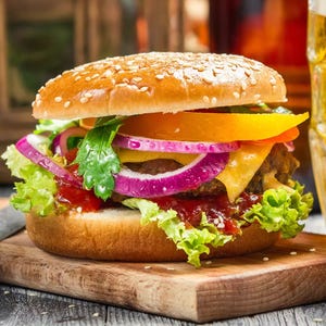 A juicy cheeseburger with a sesame seed bun, lettuce, tomato, cheddar cheese, red onions, and a sauce, served on a wooden board.
