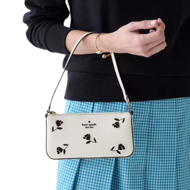 A person holds a small white handbag with a floral print and \“kate spade\“ logo, paired with a blue checkered skirt and a simple bracelet.