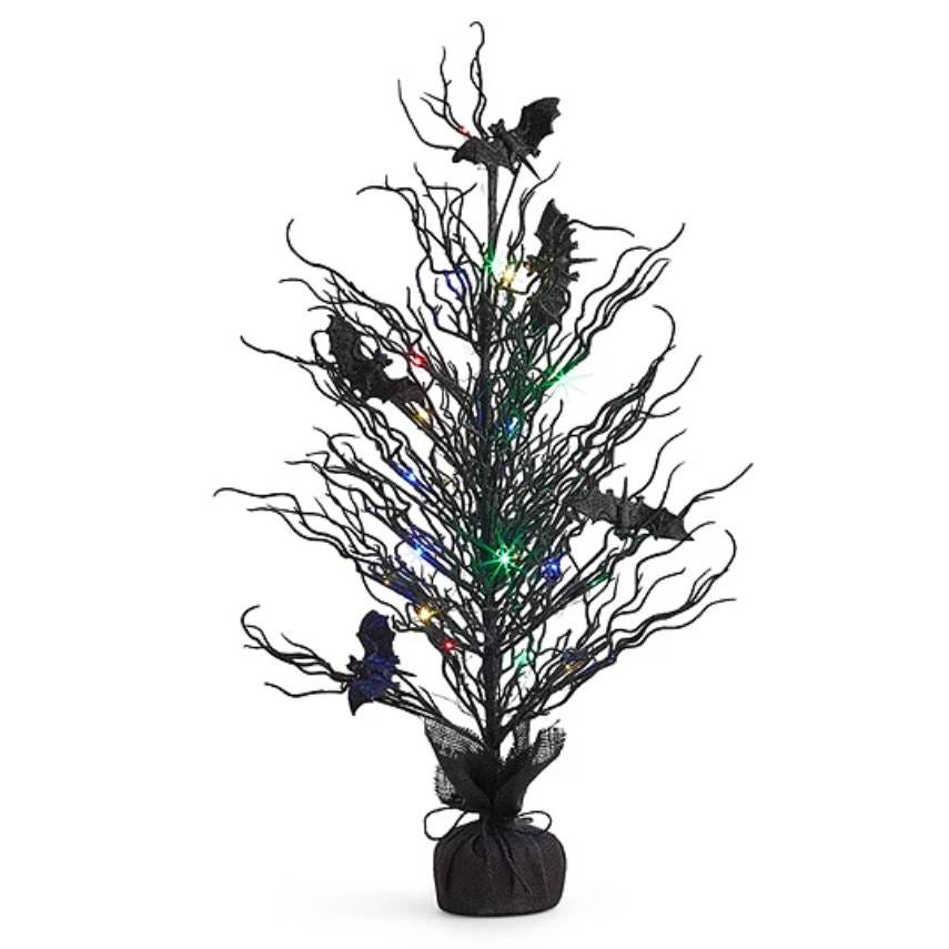 A black artificial tree with twisted branches, decorated with colorful lights and bat ornaments, set in a dark, fabric-covered base.