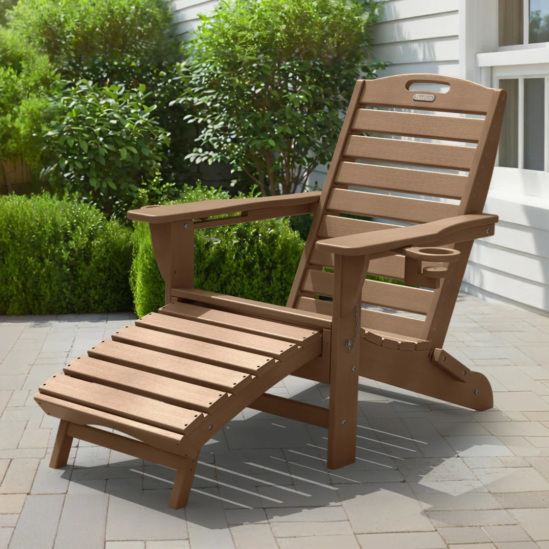 A wooden Adirondack chair with an attached footrest and a cup holder, placed on a patio near green shrubs and a white house wall.