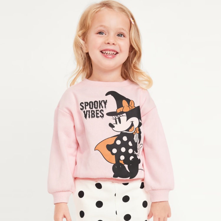 A pink sweatshirt featuring a graphic of Minnie Mouse in a black outfit with an orange bow and the text \“Spooky Vibes.\“ The sweatshirt has a crew neckline and long sleeves.