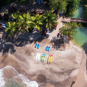 Aerial view of a beach with palm trees, blue sun loungers, two white paddleboards, and three colorful kayaks arranged on the sand near the shoreline.