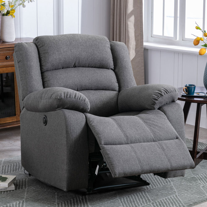 Gray overstuffed power recliner with plush cushioning, featuring a button on the side for adjusting the footrest. It has a cozy, padded design ideal for relaxation, set in a room with a side table, window, and decorative elements.
