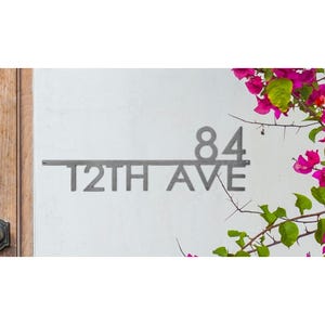 Modern house number sign displaying \“84 12TH AVE\“ in sleek metal, mounted on a wall with pink bougainvillea flowers nearby.