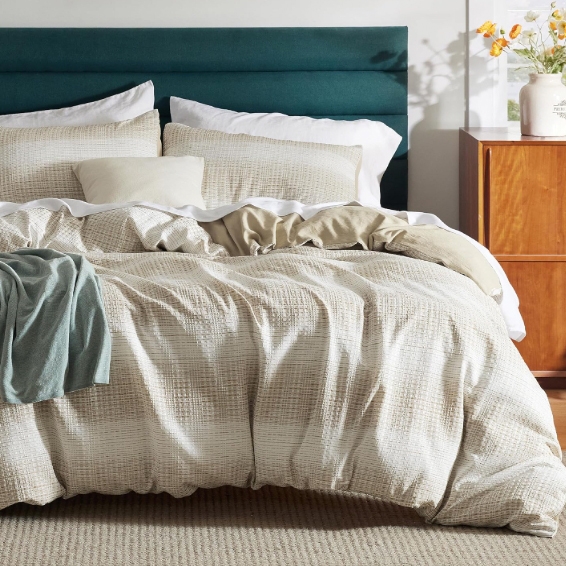 The Bedsure Queen Duvet Cover features a textured, beige fabric with a subtle grid pattern and includes matching pillow shams. It’s styled on a teal headboard with a cozy, layered look, complemented by a wooden dresser with flowers.
